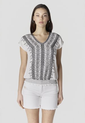 Mujer con blusa blanca de manga corta con bordado negro y pantalones cortos de mezclilla blanca desgastada, de pie contra un fondo claro y liso.