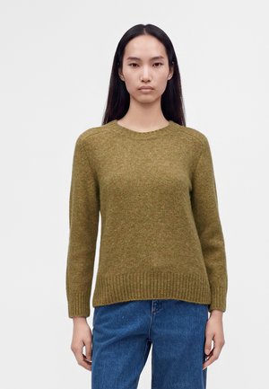 Jeune femme aux longs cheveux foncés portant un pull vert olive et un jean bleu, debout devant un fond clair uni.