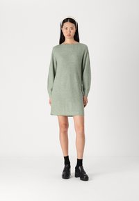 Robe tricotée vert clair avec manches longues, décolleté bateau et coupe droite. Le modèle porte des chaussettes noires et des chaussures noires épaisses.