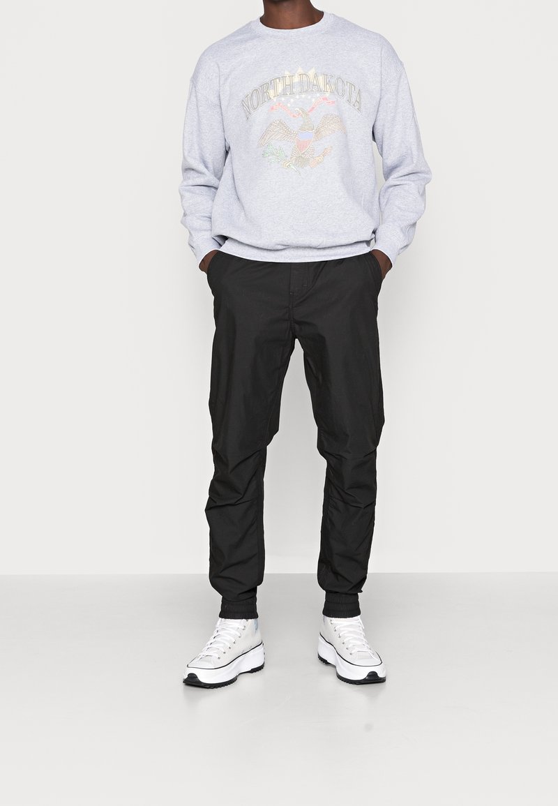 Homme portant un sweat-shirt North Dakota gris clair, un pantalon noir fuselé et des baskets blanches montantes, debout les mains dans les poches.