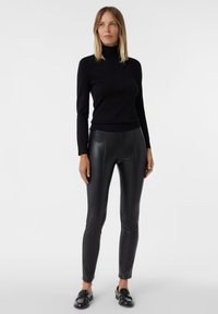 Schwarz turtleneck Pullover mit langen Ärmeln, kombiniert mit schwarzen Lederleggings. Beide Kleidungsstücke haben eine glatte Textur und einen figurbetonten Schnitt. Schwarze Mokassins vervollständigen den Look.