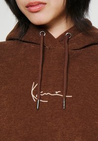 Sudadera con capucha marrón de textura suave, con un logo blanco bordado en el pecho y herrajes plateados en los cordones.