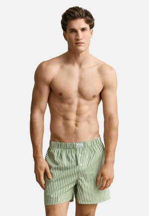 Hemdloser Mann, der grün-weiß gestreifte Boxer-Shorts mit vertikalen Streifen und Frontknöpfen trägt, steht vor einem einfarbigen hellen Hintergrund.