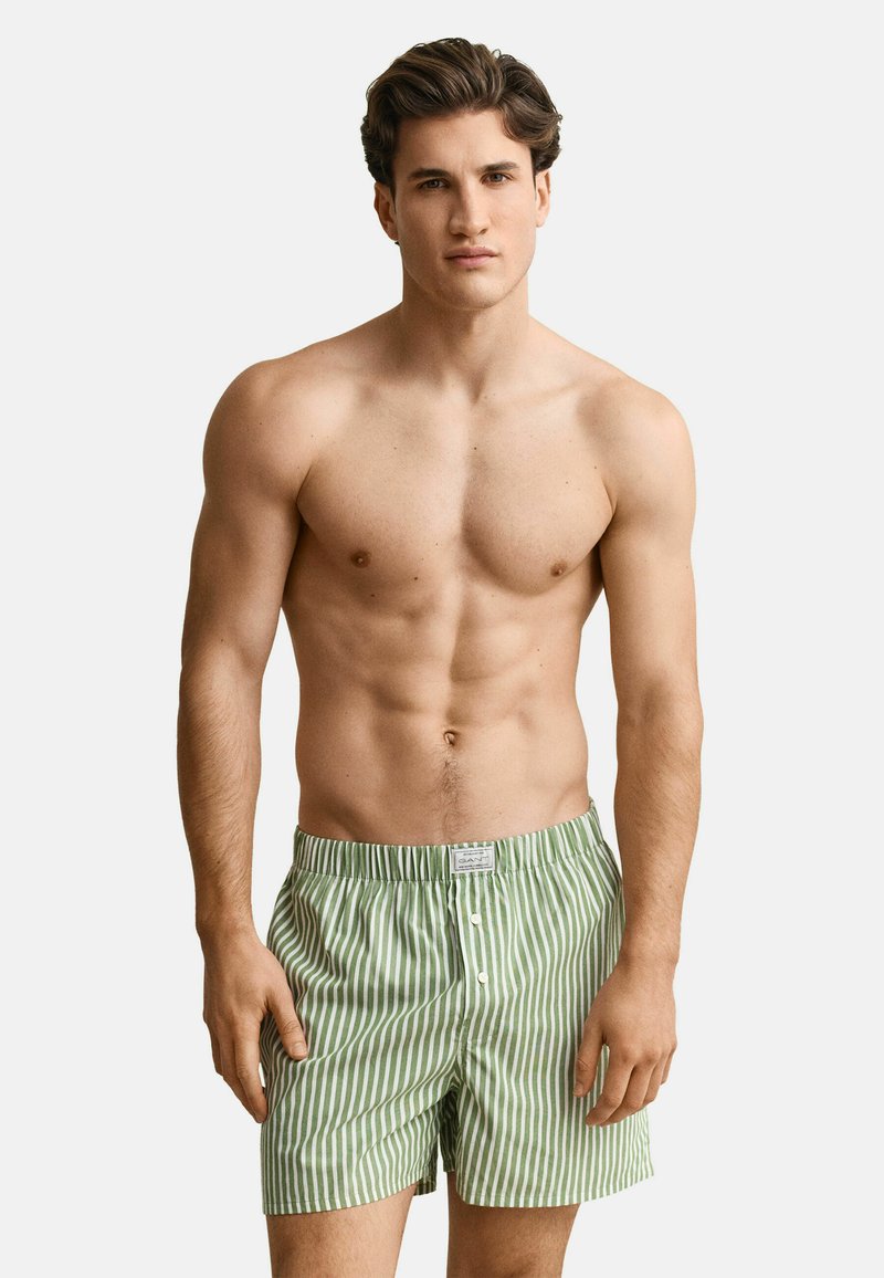 Homme torse nu portant un boxer à rayures verticales vertes et blanches avec des boutons à l'avant, debout devant un fond clair uni.
