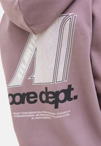 Gros plan sur un sweat à capuche mauve avec un grand motif blanc et argent affichant le texte "ATLANTIC" et "core dept." de manière bien visible.
