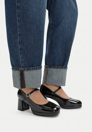 Czarne lakierowane czółenka Mary Jane z zaokrąglonym noskiem, paskiem wokół kostki i masywnym słupkowym obcasem, zestawione z krótkimi niebieskimi jeansami.