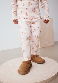 Laps kannab roosat lillemustriga soonikutoppi ja sobivaid pükse, millele on kombineeritud pruunid seemisnahast libisevad jalanõud, seistes ümmargusel platvormil.