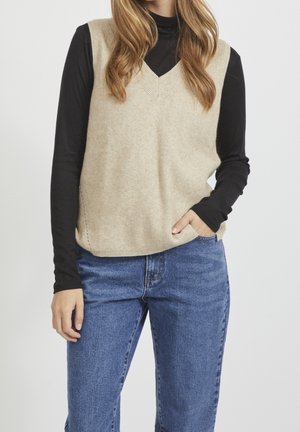 Femme portant un gilet en tricot beige sans manches sur un haut noir à manches longues et un jean bleu, une main dans la poche sur fond uni.