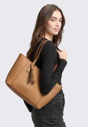 Giovane donna con lunghi capelli castani che indossa una maglia nera e jeans scuri, con una grande borsa tote in pelle color cuoio sulla spalla.