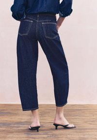 Ciemnoniebieskie dżinsowe kuloty o luźnym kroju, skróconych nogawkach i kieszeniach z tyłu. Noszone z czarnymi sandałami na obcasie na drewnianej podłodze.