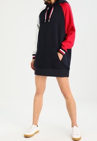 Marineblaue Hoodie-Kleid mit roten und weißen Raglanärmeln. Mit Fronttasche, gestreiften Bündchen und Kordeln ausgestattet. Aus weichem Stoff gefertigt.
