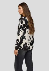 Pullover in Creme mit großem schwarzem Blumenmuster, aus strukturiertem Stoff gefertigt. Hat lange Ärmel und eine lockere Passform, von der Seite betrachtet.