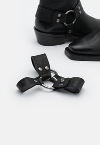 Botas de cuero negras con un acabado texturizado, que presentan herrajes metálicos en tono plateado y una correa con un anillo. El diseño de la bota es elegante y puntiagudo.