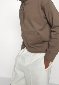 Brown hoodie with a drawstring, kangaroo pocket, and ribbed cuffs, paired with white pleated shorts. Smooth fabric texture, minimalistic design.