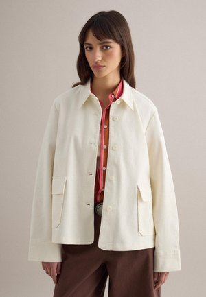 Donna con capelli castano lisci, che indossa una giacca color avorio con bottoni sopra una camicia rossa a righe e pantaloni marroni, in piedi contro uno sfondo semplice.