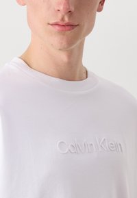 White cotton t-shirt with a crew neck, featuring raised embroidered "Calvin Klein" logo in the center. Smooth texture and minimal design.