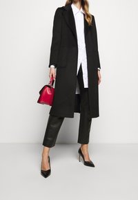 Mujer vestida con un abrigo largo negro, camisa blanca de botones, pantalones cortos negros y tacones altos negros, sosteniendo un pequeño bolso rojo.