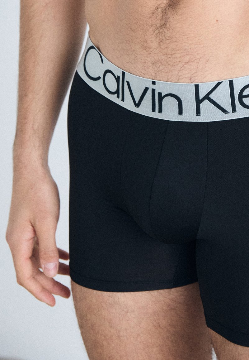 Boxer-briefs noirs avec une ceinture élastique grise portant la marque "Calvin Klein" en noir. Tissu lisse et design ajusté.