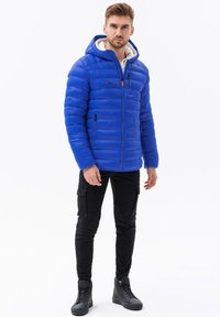 Giacca puffer blu con cappuccio, caratterizzata da un design a coste orizzontali, zip frontale e due tasche laterali, indossata con pantaloni cargo neri e stivali.