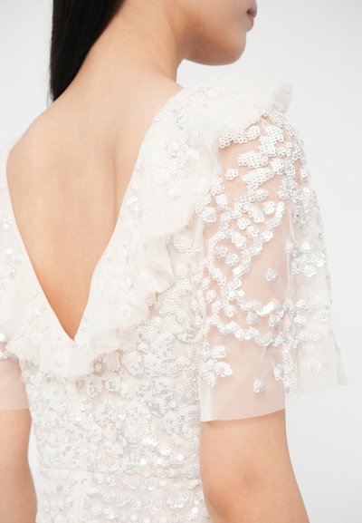 Rochie albă cu mâneci transparente cu paiete și accente cu volane. Spatele prezintă un decolteu adânc în V, evidențiind mărgele lucrate detaliat.