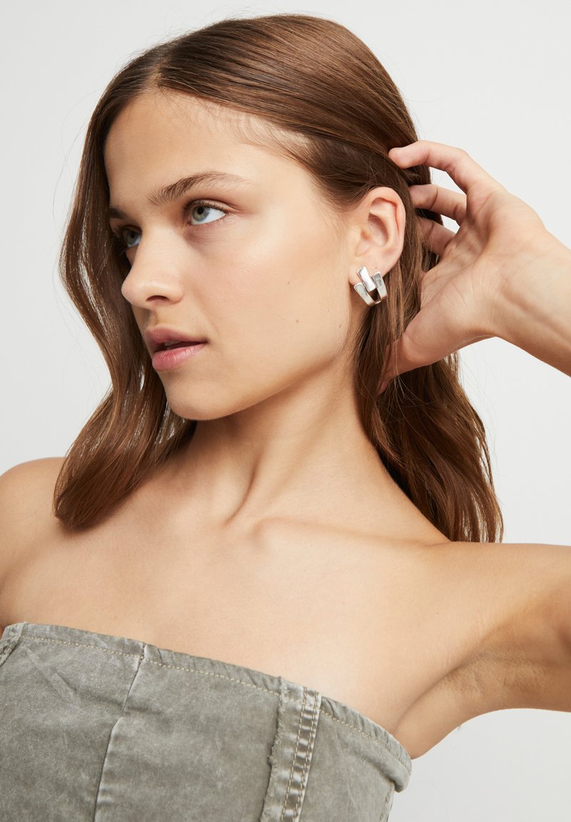 Pendientes de aro plateados con un diseño en capas que reflejan la luz, combinados con un top gris sin tirantes. Cabello peinado en ondas sueltas.