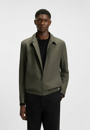 Olive green bomber jacket with a pointed collar, front zipper, and elastic hem. Worn over a black top, featuring smooth, textured fabric.