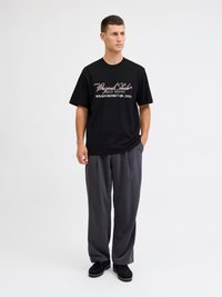 Mann steht mit den Händen in den Taschen, trägt ein schwarzes T-Shirt mit Grafik, lockere graue Hose und schwarze Schuhe vor einem schlichten weißen Hintergrund.