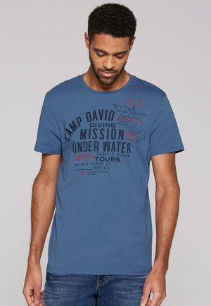 Mann trägt ein blaues Grafik-T-Shirt mit Text zum Thema Tauchen und Ozean, steht vor einem einfachen hellgrauen Hintergrund.