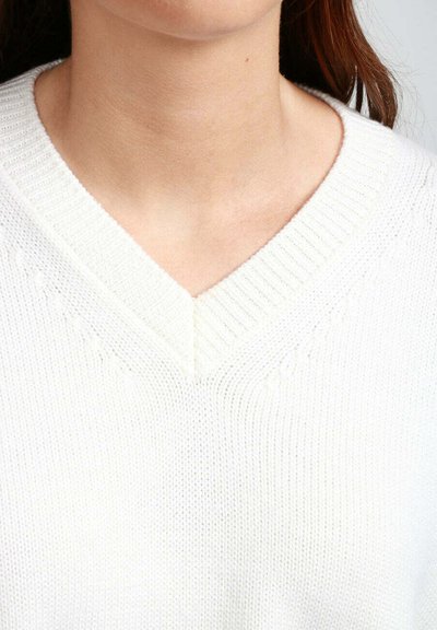 Primo piano di una persona che indossa un maglione bianco a maglia con scollo a V, che mostra il neckline e la parte superiore del torace, con un tono di pelle chiaro e capelli castani.