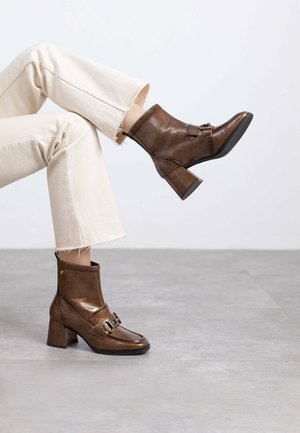 Botas de cuero marrón hasta el tobillo con un acabado brillante, punta redonda, tacón ancho y detalle decorativo de hebilla. Llevadas con jeans claros deshilachados.