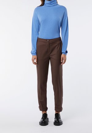 Henkilö, joka pukeutunut siniseen poolopaitaan, ruskeisiin käärittyihin housuihin ja mustiin nauhallisiin kenkiin, seisoo valkoisella taustalla.