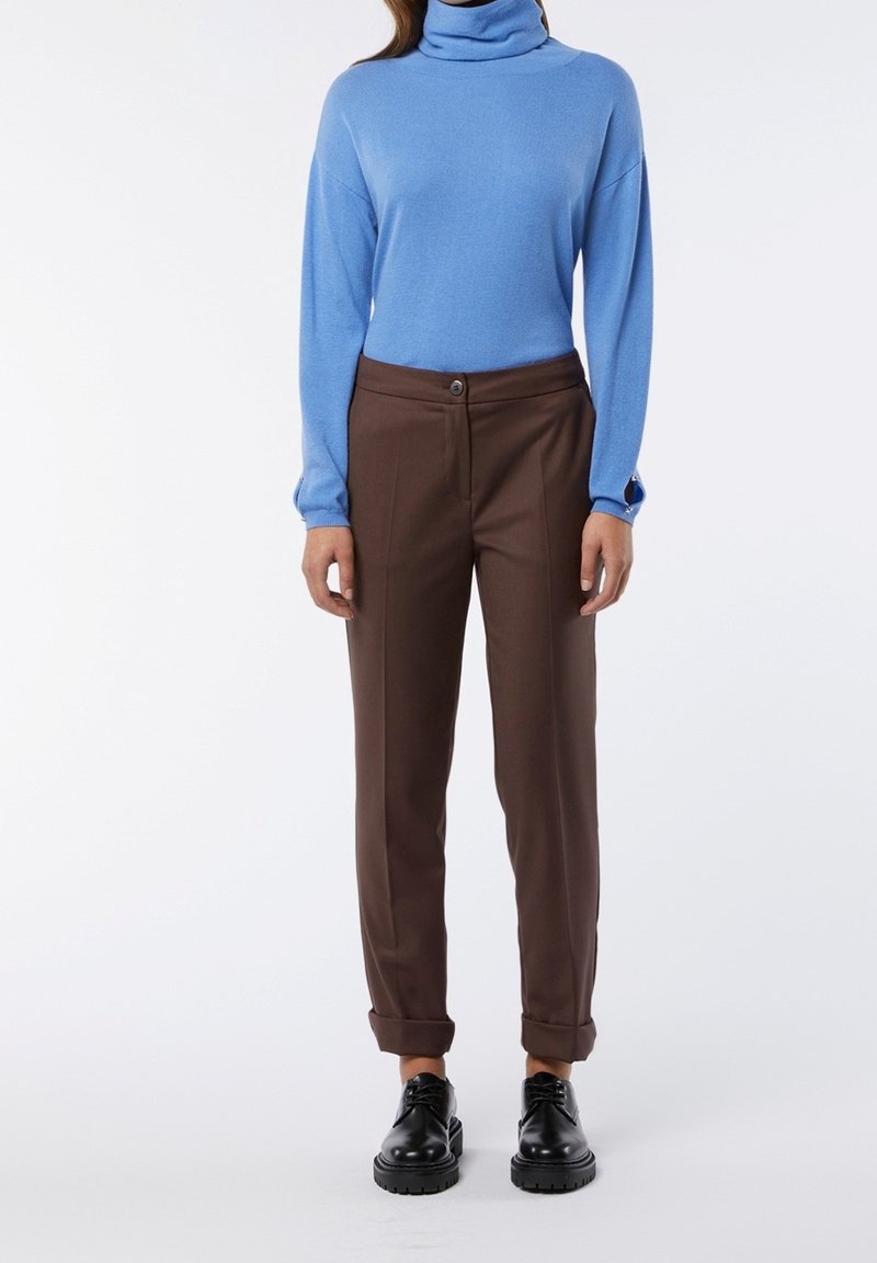 Personne portant un pull à col roulé bleu, un pantalon marron avec revers et des chaussures noires à lacets, debout sur un fond blanc.
