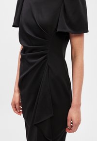 Melnā kleita ar nopludinātu dizainu, kas izgatavota no mīksta auduma, ar smalku tekstūru un diegu detaļām sānos. Īsas piedurknes ar noapaļotiem galiem.