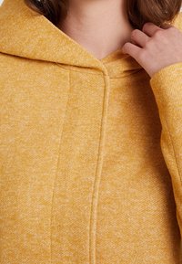 Gul hættetrøje i tekstureret stof med en blød finish. Har en stor hætte og en detalje med søm foran, der viser et lyst, plettet mønster.