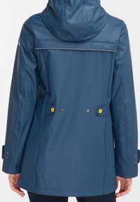 Tummansininen hupullinen sadetakki, jossa on säädettävät vyötärönauhat ja keltaiset nepparit, henkilö näyttää takaa.