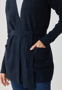 Námornícky modrý kardigán vyrobený z jemnej textílie, s opaskom v páse, rebrovanými manžetami a dvoma prednými vreckami. S jemným textúrovým detailom po celej ploche.