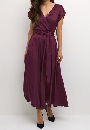Femme portant une robe portefeuille sans manches d'un violet profond avec une ceinture à la taille, associée à des escarpins noirs à bout pointu sur un fond uni.