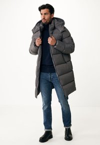 Abrigo acolchado gris con capucha, que presenta un diseño acolchado y bolsillos con cremallera, combinado con jeans oscuros y botas negras. Textura suave, hasta la rodilla.