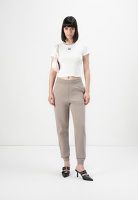 Vrouw met steil donker haar, gekleed in een witte cropped t-shirt, beige joggers, zwarte hoge muiltjes, en met een lichtroze schoudertas.