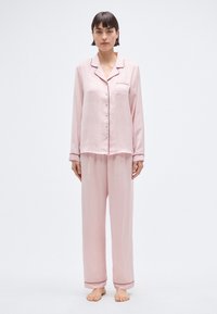 Ensemble de pyjama rose pâle en tissu lisse. Chemise à boutons avec poche et passepoil noir. Pantalon ample avec ourlets retroussés.