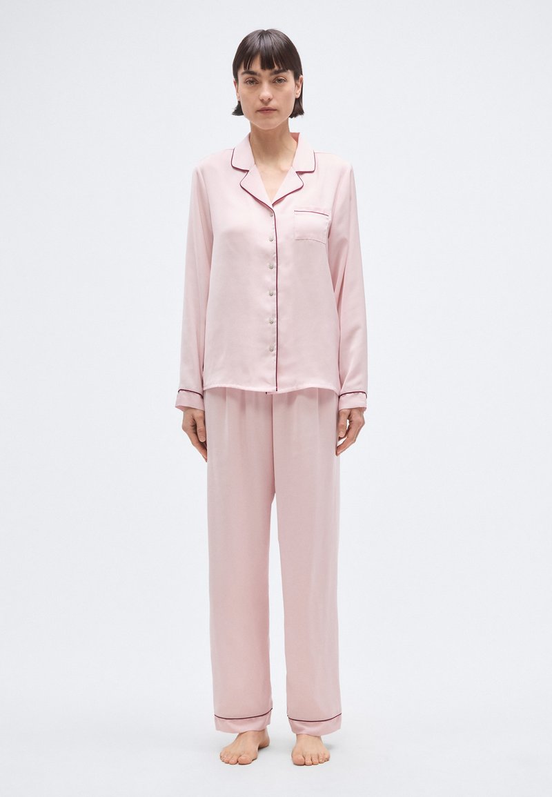 Ensemble de pyjama rose pâle en tissu lisse. Chemise à boutons avec poche et passepoil noir. Pantalon ample avec ourlets retroussés.