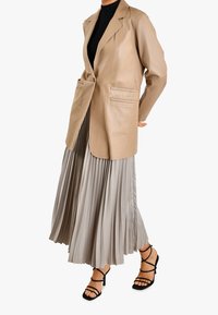 Beżowa skórzana marynarka z klapami i pojedynczym zapięciem na guzik, zestawiona z plisowaną srebrną spódnicą oraz czarnymi sandałkami na wysokim obcasie z paskami.