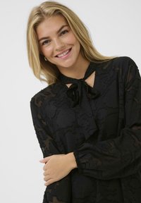 Bluză neagră din chiffon cu textură florală, guler cu fundă și mâneci lungi cu manșete gathered. Materialul transparent adaugă o apariție delicată.