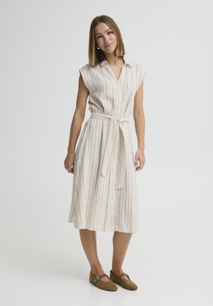 Nő ujjatlan, övvel megkötött, gombos, függőleges csíkos ruhában, barna Mary Jane lapos cipőben, egyszínű háttér előtt áll.