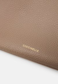 Brun læderpung med en struktureret overflade, der har et guld "Coccinelle" logo præget på forsiden. Enkel, rektangulær design.