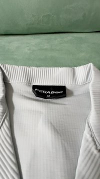 Biely textilný golier košele s ribbed textúrou, s čiernou etiketou, na ktorej je napísané "PEGADOR" a veľkosť "M." Svetlé pozadie.
