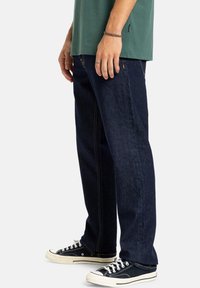 Tmavomodré riflové nohavice so strihom rovného nohu a kontrastným prešívaním, skombinované s čiernymi nízkymi teniskami.