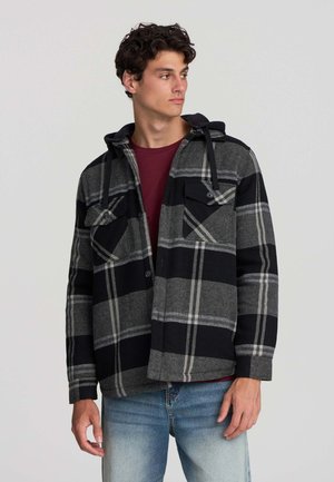 Giacca in flanella a quadri nei toni del grigio e del nero con cappuccio, dotata di due tasche sul petto e chiusura a bottoni. Indossata sopra una camicia color bordeaux.