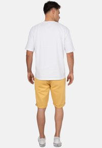 Witte katoen t-shirt met korte mouwen en gele shorts, beide van katoen. De shorts hebben opgerolde manchetten en worden gedragen met witte sneakers. Gezien vanaf de achterkant.