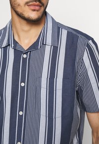 Camicia a maniche corte a righe blu navy e azzurro, con chiusura a bottoni, taschino sul lato sinistro del petto e colletto.